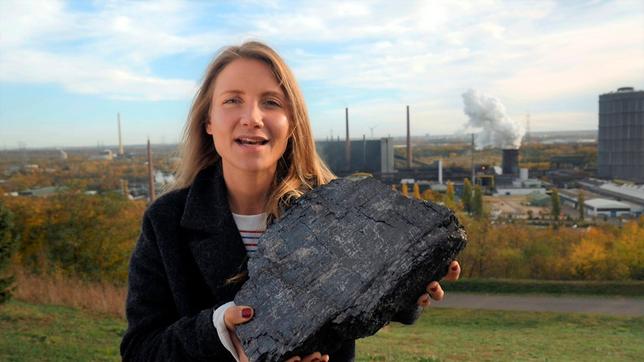 Jana steht auf einem Hügel vor der Kokerei Prosper im Ruhrgebiet. Sie hält ein großes Stück Steinkohle in der Hand.