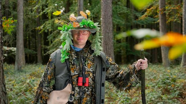 Robert steht im Tarnoutfit im Wald. Bei sich trägt er Outdoor Ausrüstung wie Kompass, Fernglas und Wanderstock.