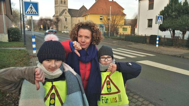 Gesa gemeinsam mit Linus und Lena an einem Zebrastreifen in der Stadt Reelsen. Alle machen den Daumen nach unten.