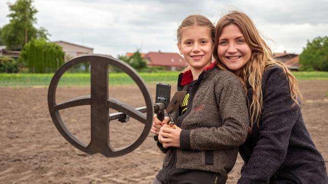 Jana kniet neben Eleni auf einem Feld und hält einen Metalldetektor in der Hand