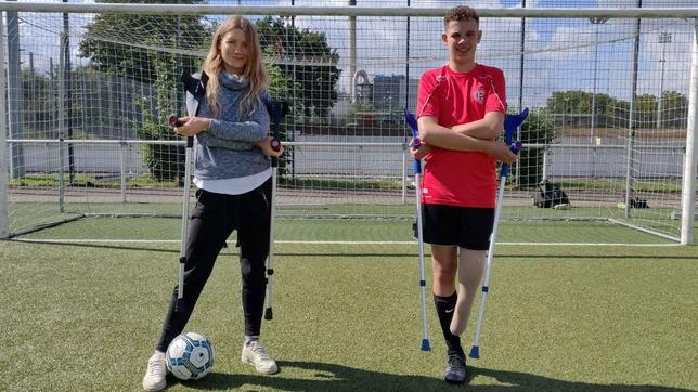 Reporterin Jana und der 14-Jährige Jamie stehen mit Krücken in den Händen vor einem Fußballtor. Jamie ist Amputierten-Fußballer, ihm fehlt ein Bein.