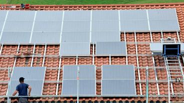 Ein Arbeiter steht auf einem Gerüst vor einem Hausdach um Solarpanel zu installieren. 