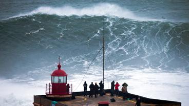 Personen schauen einem Surfer zu, der vor Nazaré auf einer riesigen Welle reitet.