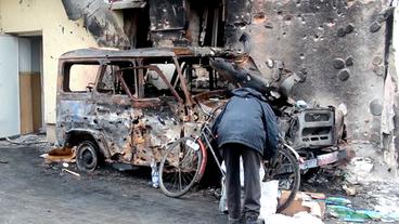 Mann mit Fahrrad vor einem zerstörten Auto.