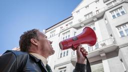 Hubert steht vor einem Haus und spricht ins Megaphon. 