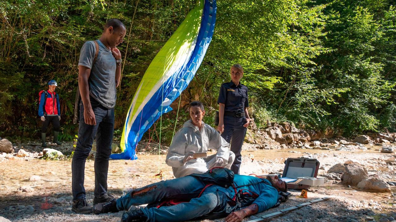 Der Profi-Paraglider Mike Bischoff ist in den Tod gestürzt. War es kein Unfall? Die Ermittlungen der Kommissare bestätigen zunehmend diese These. V.l.n.r. Jerry Paulsen (Peter Marton), Pathologin Sonja Bitterling (Genoveva Mayer), Caro Reiser (Sarah Thonig) und Komparsen